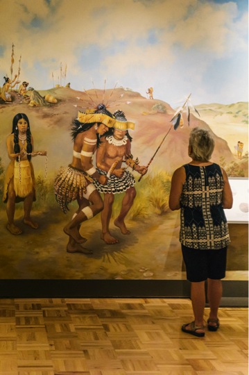Shellmound exhibit: A mural, painted by Ann Thiermann in consultation with local tribal members and historians, depicts an imagined scene in a Huchuin Ohlone village. Elevated above the Bayâ€™s tides, the Shellmounds depicted in the mural served as sacred burial sites, and as settings for homes and ceremonies. Photo by Shaun Roberts, courtesy Oakland Museum of California